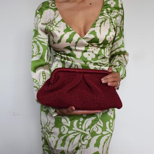 Puede incluir: Un clutch burdeos de ganchillo con un diseño fruncido. El clutch se sostiene sobre un fondo de un vestido con estampado floral verde y blanco. El bolso tiene forma rectangular con la parte superior redondeada.