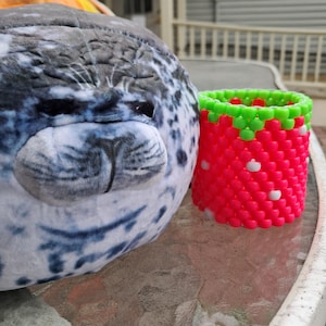 May include: A soft seal toy with a grey and white spotted design, beside a handmade beaded strawberry-shaped container. The container is red with green and white details, resembling a strawberry.