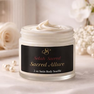 May include: A glass jar filled with white body soufflé. The jar has a black label with gold and burgundy text that reads "Selah Sacred Sacred Allure 2 oz Satin Body Soufflé". The jar is sitting on a white surface.