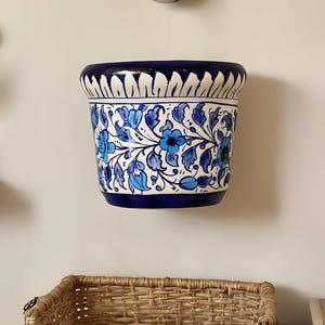 Puede incluir: Jardinera de pared de cerámica azul y blanca con diseño floral. La jardinera tiene un borde azul oscuro y un interior blanco. El estampado floral presenta flores y hojas azules. La jardinera tiene unos 20 cm de diámetro.