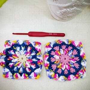 May include: Two colorful, square crochet pieces with a floral design, featuring shades of blue, pink, yellow, and white. A red crochet hook, marked 7/0 4.0mm, lies between them. The background is a neutral, textured surface.