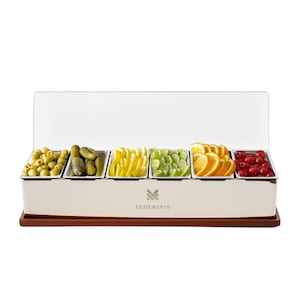 May include: A white serving tray with six compartments, each filled with different foods. The tray holds olives, pickles, sliced lemons, limes, oranges, and cherries. The tray sits on a wooden base and has a white lid. The brand name "SEDERINIS" is printed on the front.