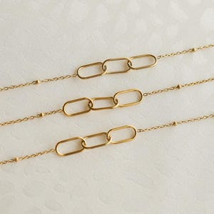 May include: Three gold-toned necklaces arranged horizontally, each featuring a delicate chain with small evenly spaced beads and a central focal point consisting of three elongated oval links connected in a row, creating a uniform design.