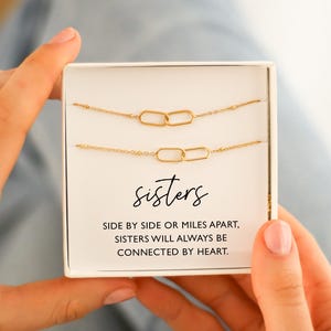 Könnte beinhalten: Zwei goldfarbene Kettenarmbänder mit ineinandergreifenden ovalen Gliedern werden in einer weißen Schachtel präsentiert. Das Wort "sisters" ist in Schreibschrift unter den Armbändern geschrieben. Der Text darunter lautet: "SIDE BY SIDE OR MILES APART, SISTERS WILL ALWAYS BE CONNECTED BY HEART."