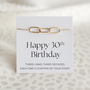 Op de afbeelding: Een goudkleurige kettingarmband met drie verbonden ovale bedels, gepresenteerd op een witte kaart. De kaart leest "Happy 30th Birthday" met de tekst "THREE LINKS, THREE DECADES, EACH ONE A CHAPTER OF YOUR STORY."