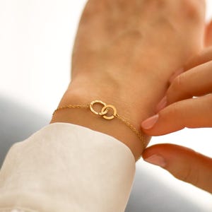 May include: A gold bracelet with two linked circles. The bracelet is attached to a fine chain and is worn on a wrist. The circles have a textured design. The bracelet is a piece of jewellery.