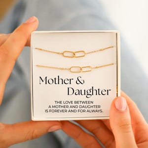 May include: A gold-coloured necklace set for a mother and daughter, presented in a white box. The necklaces feature linked oval pendants. The box has the words "Mother & Daughter" and a message about the love between them.