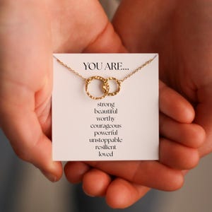 Könnte beinhalten: Eine goldfarbene Halskette mit zwei ineinandergreifenden Kreisen, präsentiert auf einer weißen Karte. Auf der Karte steht "YOU ARE..." gefolgt von inspirierenden Worten. Die Halskette wird in offenen Händen gehalten.