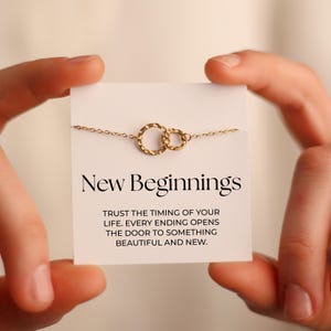 May include: A gold-coloured necklace featuring two linked circles is shown on a white card. The card reads "New Beginnings" with an inspirational quote. The necklace is held between two hands.