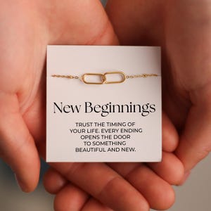 May include: A gold-toned chain bracelet featuring two interlocking oval links centered on a white display card. The card displays the text New Beginnings followed by the phrase Trust the timing of your life. Every ending opens the door to something beautiful and new.