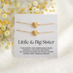 Könnte beinhalten: Zwei goldfarbene Armbänder mit Herzanhängern auf einer weißen Karte mit dem Text "Little & Big Sister". Die Karte enthält auch den Text "TWO HEARTS, ONE UNBREAKABLE BOND. GROWING, CHANGING, BECOMING - YET ALWAYS HOLDING ON TO EACH OTHER. SISTERS FIRST, FRIENDS FOREVER."