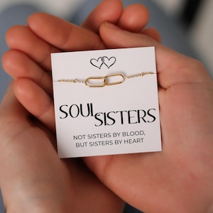 Puede incluir: Una pulsera de cadena dorada con dos eslabones ovalados entrelazados, presentada en una tarjeta blanca. La tarjeta presenta las palabras "SOUL SISTERS" y la frase "NOT SISTERS BY BLOOD, BUT SISTERS BY HEART" en texto negro, con dos ilustraciones de corazones.