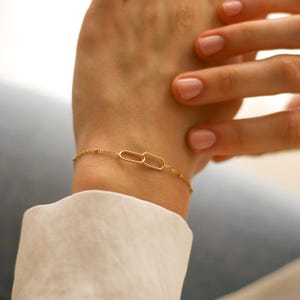 May include: A gold-coloured bracelet with a fine chain and small beads, featuring two interlocking oval links. The bracelet is displayed on a wrist, highlighting its minimalist design. A simple and elegant piece of jewellery.