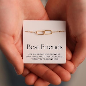 Könnte beinhalten: Ein goldfarbenes Kettenarmband mit zwei verbundenen ovalen Anhängern, präsentiert auf einer weißen Karte. Die Karte trägt die Aufschrift "Best Friends" mit einer Botschaft über Freundschaft. Das Armband wird in offenen Händen gehalten.