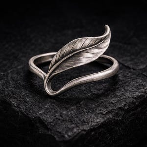 Puede incluir: Anillo plateado con un diseño detallado de hoja. La hoja está posicionada verticalmente, con la banda del anillo curvándose alrededor del dedo. El anillo se encuentra sobre un fondo oscuro y texturizado.