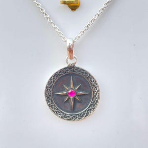 May include: A silver pendant necklace featuring a circular pendant with an eight-pointed star design and a bright pink gemstone at the center. The pendant has an ornate, swirling border and hangs from a delicate silver chain. A small gold-colored charm is visible above the pendant.