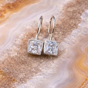 Op de afbeelding: Een paar zilveren oorhangers met vierkante, heldere edelstenen. De oorbellen hebben een eenvoudig, elegant ontwerp, met een vierkante edelsteen gezet in een zilveren montuur. De oorbellen worden gepresenteerd op een lichtbruin en wit oppervlak.