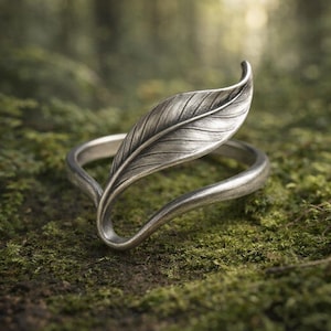 May include: A silver ring with a leaf design. The leaf detail curves over the finger, with the band forming an open shape. The ring is set against green moss, enhancing its natural aesthetic. The ring's design is reminiscent of nature.