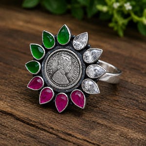 Puede incluir: Un anillo de plata con una moneda central que representa a la reina Victoria de perfil, rodeado por un halo en forma de flor de piedras preciosas en forma de lágrima en colores alternos verde, blanco transparente y rosa intenso.