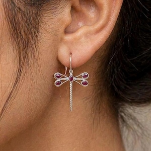 Puede incluir: Pendientes de plata con diseño de libélula y gemas de color rubí. El diseño de la libélula presenta un cuerpo y alas texturizadas, con las gemas engastadas en las alas y el cuerpo.