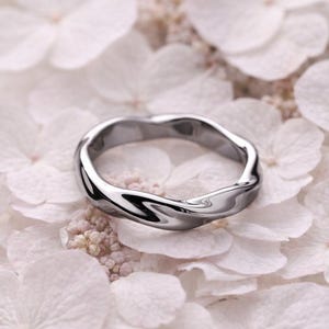 Może przedstawiać: Srebrny pierścionek o falistym wzorze, spoczywający na białych kwiatach. Powierzchnia pierścionka odbija światło, podkreślając jego gładką, wypolerowaną fakturę. Unikalny kształt pierścionka dodaje biżuterii nowoczesnego charakteru.