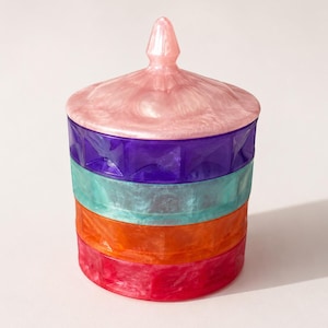 May include: A multi-coloured lidded jar with a faceted design. The jar features stacked bands in pink, purple, teal, orange, and red. The lid is pearlescent pink with a pointed finial. The jar is likely for storage or decorative purposes.