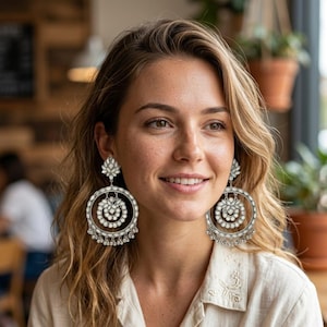 Könnte beinhalten: Silberfarbene Creolen mit floralem Design und hängenden Akzenten. Die Ohrringe haben einen großen kreisförmigen Reifen mit einem kleineren floralen Design in der Mitte und einem kleineren floralen Design oben. Getragen von einer Person.