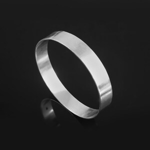 Könnte beinhalten: Ein silberfarbenes, flaches Armreif-Armband. Das Armband hat eine glatte, polierte Oberfläche und ein schlichtes, minimalistisches Design. Das Armband wird vor einem schwarzen Hintergrund mit seinem Spiegelbild gezeigt.