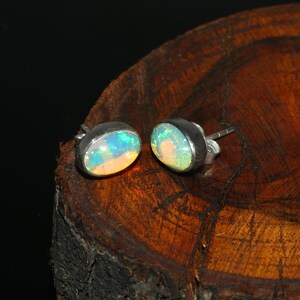 Könnte beinhalten: Ein Paar ovale Opal-Ohrstecker. Die Opale zeigen ein lebendiges Farbspiel mit Blitzen von Blau, Grün und Orange. Die Ohrringe sind in einem silberfarbenen Metall gefasst. Die Ohrringe werden auf einer dunklen Holzoberfläche präsentiert.