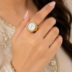Könnte beinhalten: Ein goldfarbener Ring mit integrierter Uhr, rundem weißem Zifferblatt, schwarzen Stundenmarkierungen und dem schwarzen Schriftzug BOZP QUARTZ. Die Uhr hat ein metallisch goldenes Finish, eine runde Lünette und eine kleine seitliche Krone.