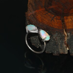 Könnte beinhalten: Ein Silberring mit zwei Opal-Edelsteinen. Einer der Opale ist rechteckig, der andere tropfenförmig. Die Opale zeigen ein Farbspiel mit schillernden Farbtönen von Blau, Rosa und Gelb. Der Ring ist vor einem dunklen Hintergrund platziert.