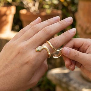 Könnte beinhalten: Ein goldfarbener Schlangenring mit einem Kopf, der mit kleinen, klaren Kristallen und grünen Akzenten verziert ist. Der Ring umschlingt den Finger in einem geschwungenen Design. Der Ring wird an einer Hand gezeigt.