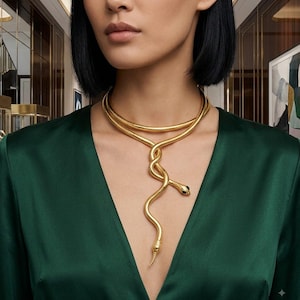 Pode incluir: Um colar tipo gargantilha dourado em forma de serpente com um design liso e enrolado que envolve o pescoço e desce. A cabeça da serpente é detalhada com pequenos olhos pretos e o corpo tem um acabamento metálico polido e reflexivo.