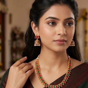 Könnte beinhalten: Goldfarbenes Schmuckset mit Halskette und passenden Ohrringen. Die Halskette und die Ohrringe sind mit abwechselnd roten und grünen tropfenförmigen Edelsteinen verziert. Die Ohrringe haben ein glockenförmiges Design mit kleinen weißen Perlen.