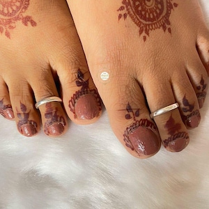 Könnte beinhalten: Nahaufnahme von zwei Füßen mit Henna-Designs und silbernen Zehenringen. Die Zehennägel sind in einem Mauve-Ton lackiert und mit aufwendigen Henna-Mustern in einem tiefbraunen Farbton verziert. Die Henna-Designs umfassen florale und geometrische Motive.