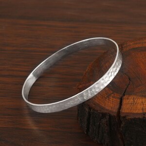 Könnte beinhalten: Ein silberfarbenes Armreif-Armband mit einer gehämmerten Textur auf der Außenseite. Das Armband ist kreisförmig und liegt auf einer dunklen Holzoberfläche. Das Armband ist ein schlichtes, elegantes Schmuckstück.