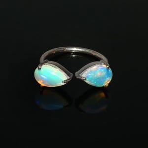 Könnte beinhalten: Ein silberner Ring mit zwei tropfenförmigen Opalen. Die Opale zeigen schillernde Farben in Blau, Grün und Orange. Der Ring ist vor einem schwarzen Hintergrund platziert, der die Edelsteine hervorhebt.