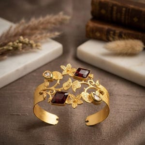 May include: A gold cuff bracelet featuring an intricate openwork design of leaves and flowers. The piece is accented with two square-cut deep red garnet stones and two smaller, clear, teardrop-shaped stones set within the metal foliage.