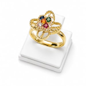 Könnte beinhalten: Ein goldener Ring mit einer blumenförmigen Fassung und einem perlenartigen Rand. Die Mitte der Blume ist mit fünf runden Edelsteinen in Blau, Orange, Grün, Weiß und Rot verziert, die auf einem polierten Goldring mit glatter Oberfläche sitzen.