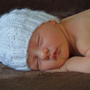May include: A sleeping baby wearing a white knitted hat. The baby is lying on a brown furry surface.