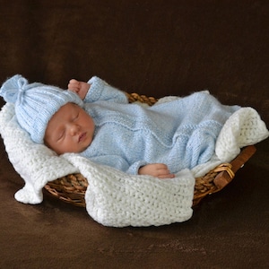 May include: A newborn baby sleeping in a wicker basket, wearing a light blue knitted hat and matching knitted outfit.