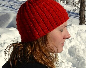 Puede incluir: Una mujer con un gorro de punto rojo con un patrón de punto de cable. El gorro está tirado sobre sus orejas y frente. Está de pie en un paisaje nevado.