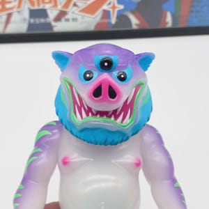 May include: A colorful, stylized vinyl figure of a creature with a pig-like snout, three eyes, and sharp teeth. The figure has a purple and blue head, a blue neck, and a translucent white body with green accents. The figure is a collectible toy.