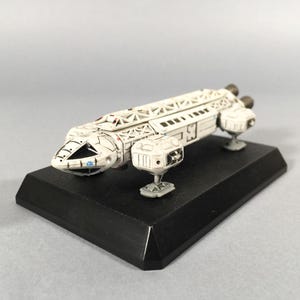 May include: A white and gray model spaceship with a detailed design, mounted on a black rectangular base. The spaceship features multiple compartments, engines, and a detailed front section. The model is approximately 10 inches long.