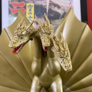 May include: A golden, three-headed dragon figurine with red mouths and detailed scales. The figure has a large, fan-shaped structure behind the heads. The background includes a Japanese poster.