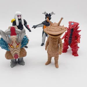 Könnte beinhalten: Eine Sammlung von fünf farbenfrohen Actionfiguren. Die Figuren umfassen eine rote und schwarze stachelige Kreatur, eine braune insektenartige Figur, eine schwarz-graue Figur, eine Figur mit einem gehörnten Kopf und eine Figur mit weißem Kopf und schwarzem Anzug.