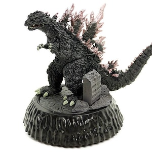 May include: A black Godzilla figure with pink dorsal fins and a long tail. The figure stands on a black base with a miniature building. The figurine is a collectible item.