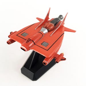 May include: A red model spaceship with black detailing and silver components, mounted on a black rectangular base. The toy features a streamlined design with multiple fins and engine details, suggesting a futuristic theme.