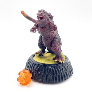 May include: A red and black Godzilla figurine with a yellow base and a separate orange atomic breath effect. The figurine is highly detailed and stands on a black, textured base.