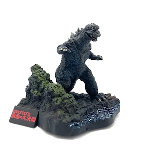 Peut inclure: Figurine de Godzilla gris foncé sur un socle rocheux avec des accents verts et bleus. La figurine est texturée et posée avec les bras tendus. Une petite base rectangulaire noire avec du texte japonais est devant.